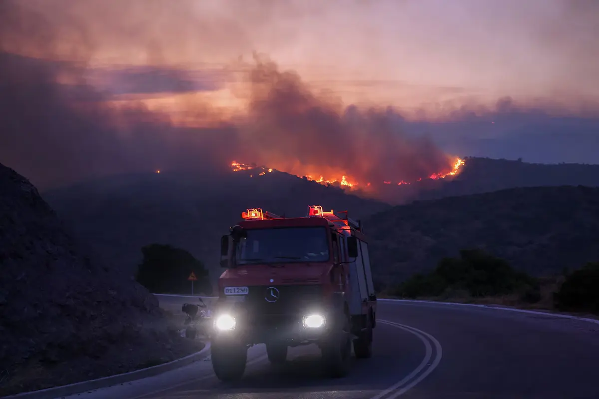 A firefighting vehicle drives down a road as flames rise from a wildfire burning on the island of Chios, Greece