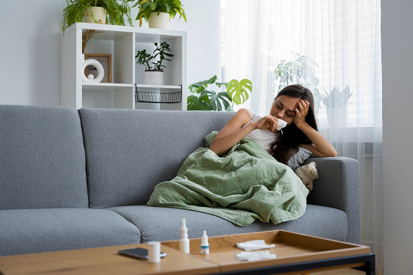 Woman sitting on a couch at home with a tissue and nasal spray, feeling unwell due to poor indoor air quality.